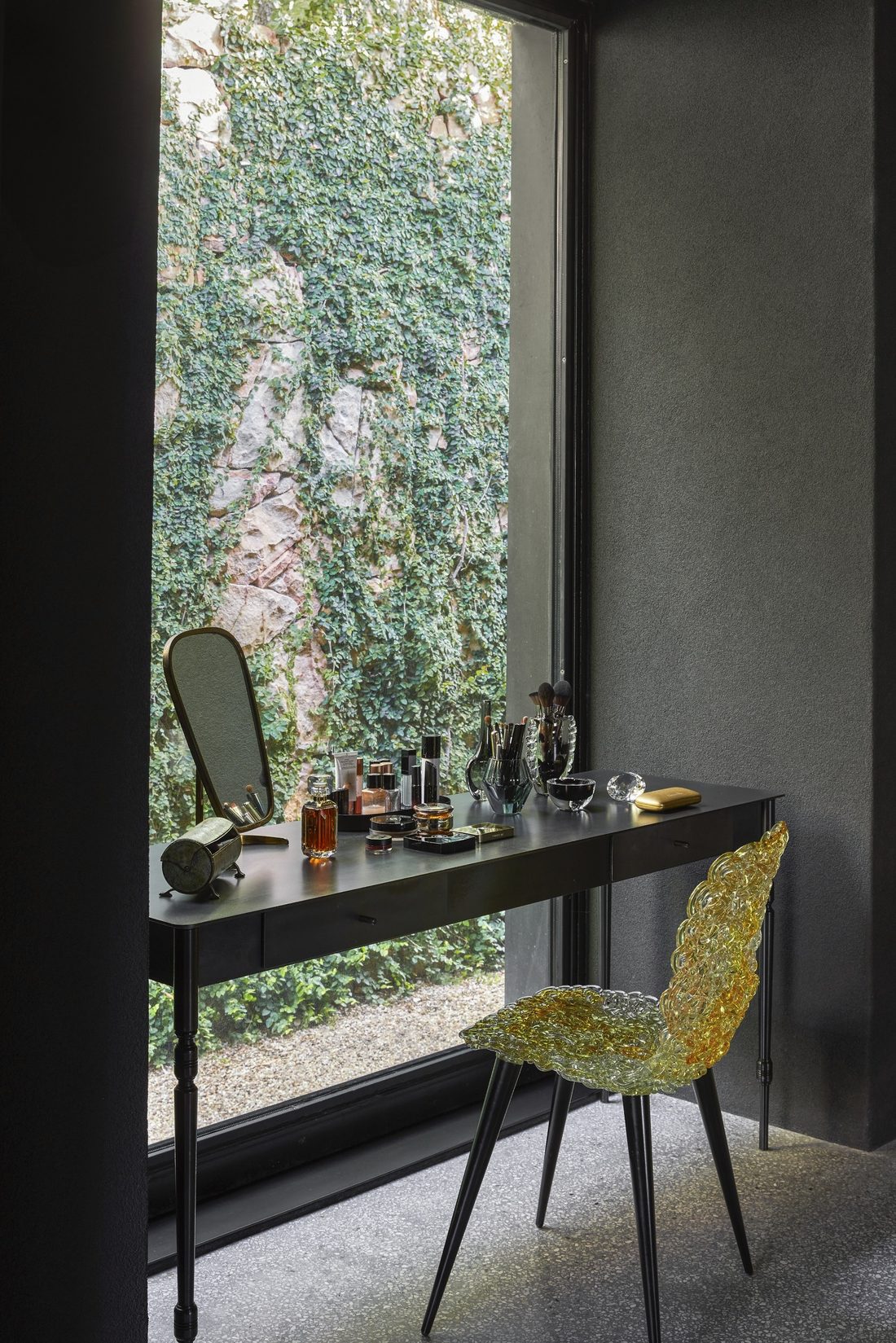 Dressing table by a tall window onto an ivy covered rock face, with a glass chair