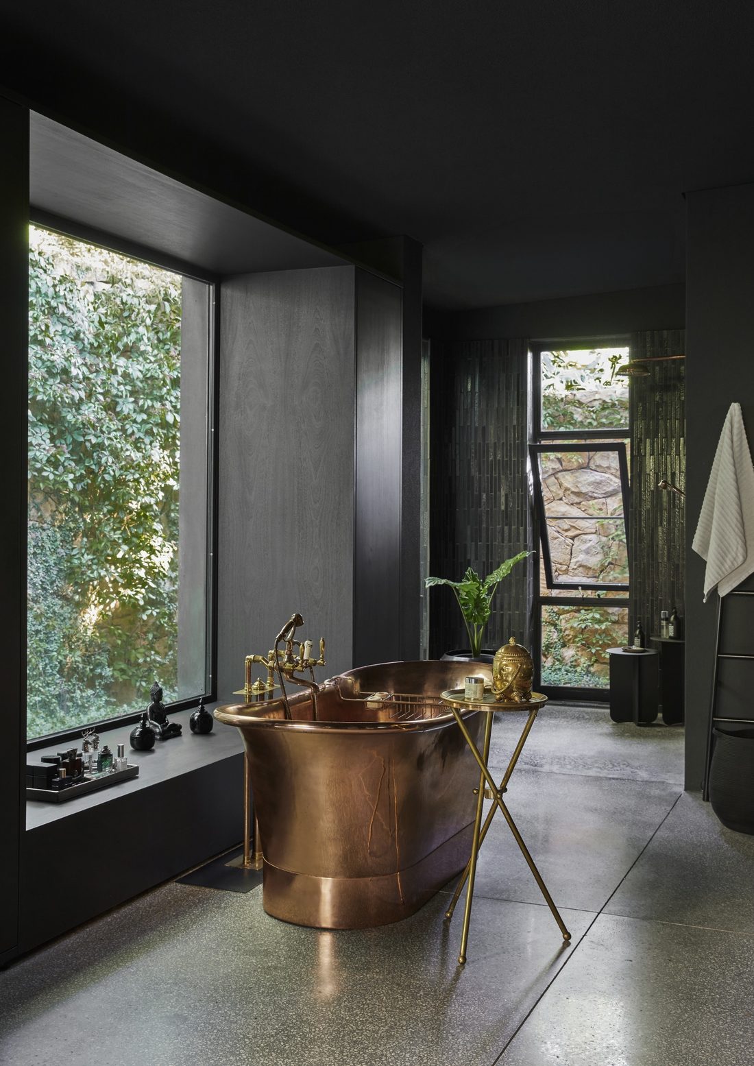 Copper bathtub beside a tall window onto an ivy covered wall, with a brass side table and a dark tiled shower beyond