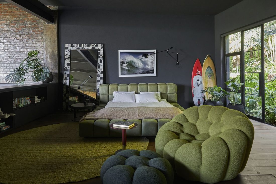 Bedroom with a green upholstered bed, a sculptural armchair and two surfboards