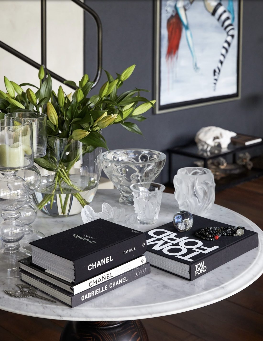 Round marble table with a vase of lilies, crystal bowls and candle holders, and stacked books