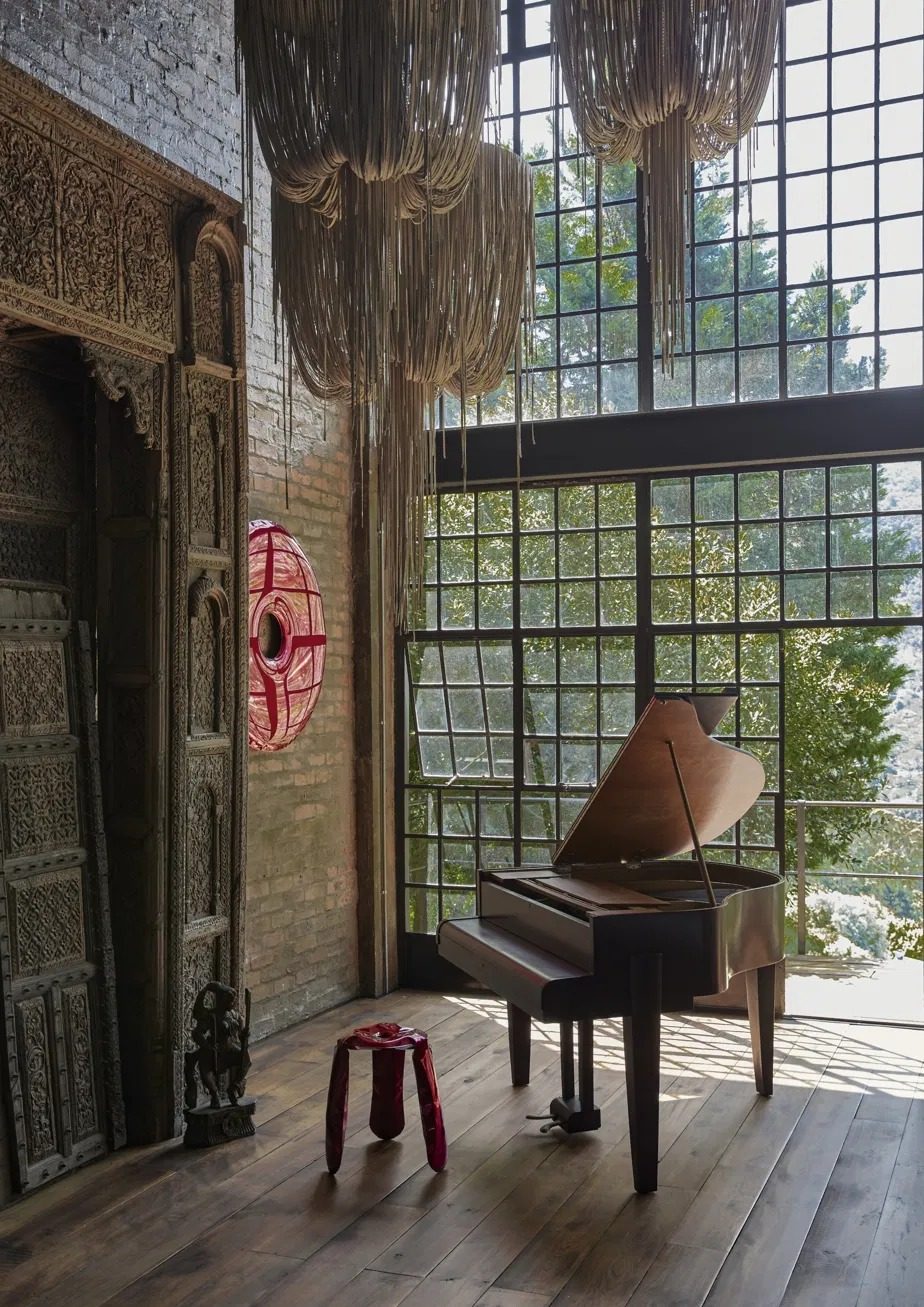 Grand piano in front of a tall steel framed window beneath fringed chandeliers, with a carved timber doorway and a red glass sculpture