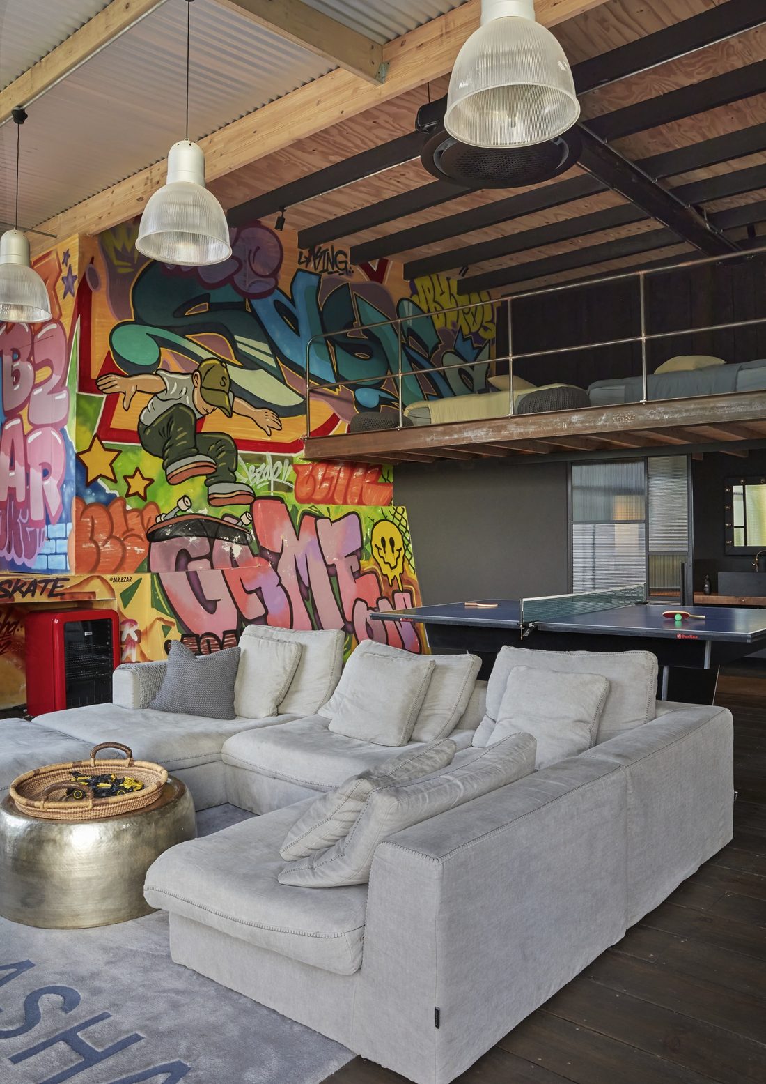 Games lounge with a graffiti mural, a grey sectional sofa, a ping pong table and a mezzanine bed above